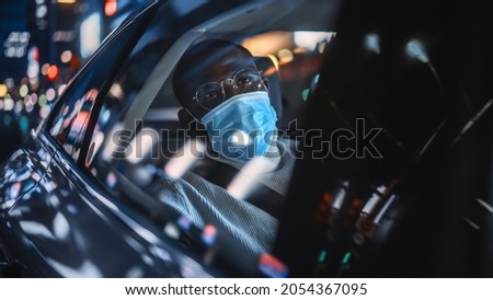 Stylish Black Man Wearing a Face Mask Commuting Home in a Backseat of a Taxi at Night. Handsome Male Passenger Looking Out of Window while in a Car in Urban City Street with Working Neon Signs.