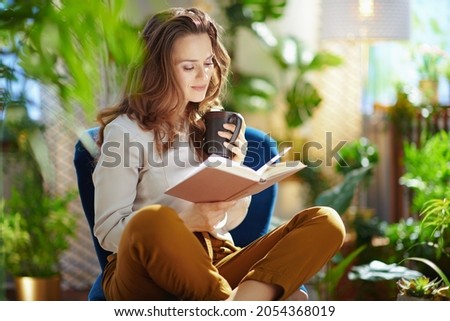Green Home. elegant woman with long wavy hair with cup of cappuccino and book in green pants and grey blouse sitting in a blue armchair at modern home in sunny day. Royalty-Free Stock Photo #2054368019
