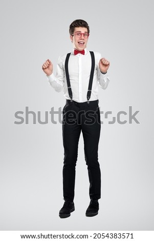 Full body of happy young man in nerdy glasses wearing white shirt with suspenders and bow tie looking at camera while standing with fists up, against gray background