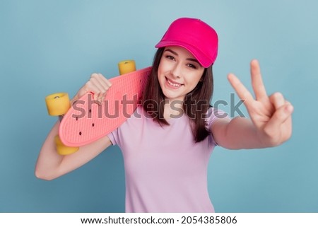 Photo of funny cute young lady wear hat holding longboard show peace v-sign isolated turquoise color background