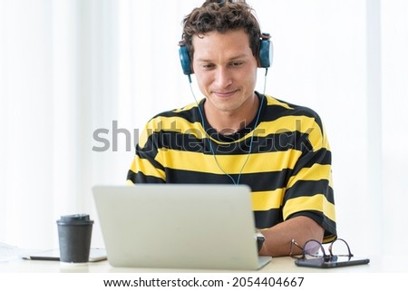 Handsome man with curly hair sitting at home playing online games with headphones and listening to music happily. 