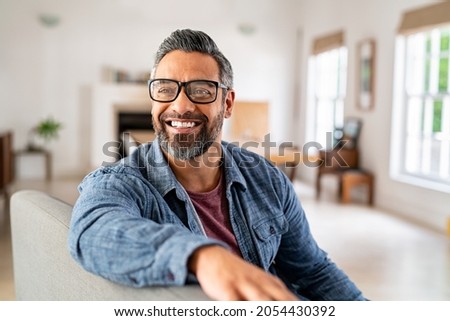 Happy mature middle eastern man wearing eyeglasses sitting on couch. Portrait of indian man relaxing at home and looking away with big smile. Mid adult guy with specs thinking about his future. Royalty-Free Stock Photo #2054430392