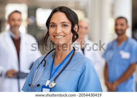 Portrait of happy young nurse in uniform with healthcare team in background. Successful team of doctor and nurses smiling. Beautiful and satisfied healthcare worker in private clinic looking at camera Royalty-Free Stock Photo #2054430419