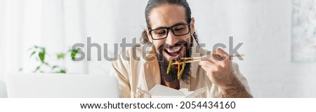 positive hispanic freelancer in eyeglasses eating chinese noodles near blurred laptop, banner