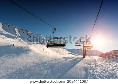Ski lift empty ropeway on hilghland alpine mountain winter resort on bright sunny evening . Ski chairlift cable way with people enjoy skiing and snowboarding. Sunset sky backlit shining on background Royalty-Free Stock Photo #2054441087