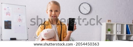 smiling girl with toy bunny holding cellphone with blank screen in clinic, banner