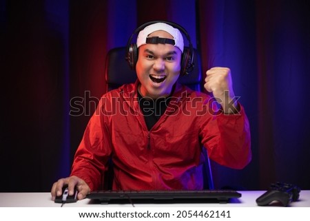 Playing video game. Young asian handsome man sitting on chair in living room. Happiness Streamer Indian man  playing game online wearing headphone in the darkroom.