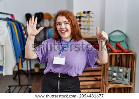 Young redhead woman working as manager at retail boutique showing and pointing up with fingers number six while smiling confident and happy. 