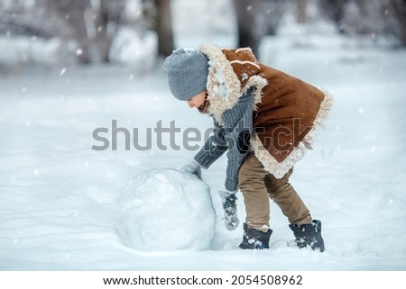 Happy boy playes on snowy winter day in the park. Child playing with snow in winter and rolling snow ball for making a snowman 
