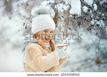 Blonde kid with cup of cocoa or hot chocolate in the winter park . Little girl holding cup with tasty marsmallow and licking