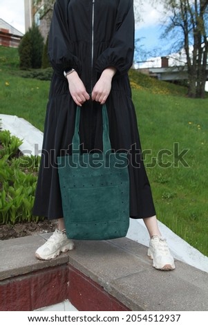 Outdoor cropped fashion portrait of woman wearing long oversize black dress, holding green leather shopper bag, posing outside over grass. Copy, empty space for text. Handbags  Fashion Accessories