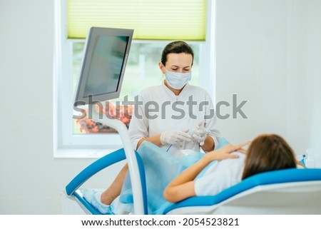 Experienced female gynecologist in lab coat, face mask and gloves holding medical vaginal speculum for examining patient. Young woman lying on gynecological chair during check up. Royalty-Free Stock Photo #2054523821