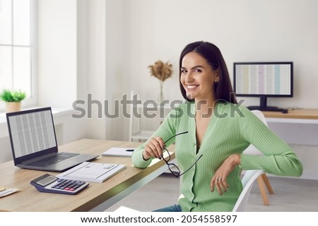 Portrait of beautiful happy female accountant sitting at her workplace in office. Smiling accountant or business analyst in casual clothes sitting in front of laptop , documents and calculator. Royalty-Free Stock Photo #2054558597