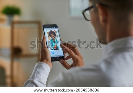 Single man flirting with beautiful woman on online dating app or website. Guy looking at mobile phone display and giving like to pretty black lady on marriage web site. Close up view over shoulder