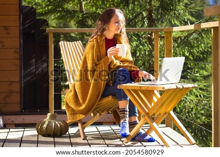 Pretty girl sitting on balcony terrace wrapped in yellow plaid holding cup of tea coffee, looking at laptop. Brunette woman works remotely, reading online. Countryside house. Wood at sunny autumn day. Royalty-Free Stock Photo #2054580929