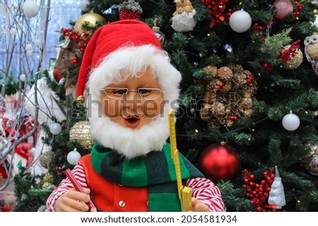 Toy Santa Claus holds a large pencil and a wooden ruler in his hands. Doll Santa Claus with glasses and a white beard near the Christmas tree. Santa Claus Doll with a construction tool for Christmas.
