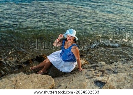 A young woman in a white skirt and a sun hat on the seashore sits on a stone admiring nature. Horizontal photo.