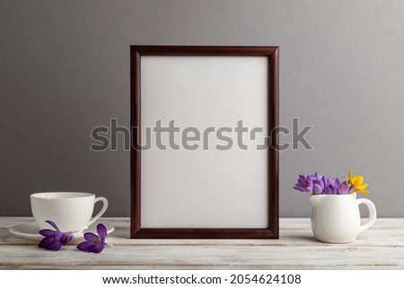 Brown wooden frame mockup with snowdrop crocus flowers and white coffee cup on gray paper background. Blank, vertical orientation, still life, copy space.