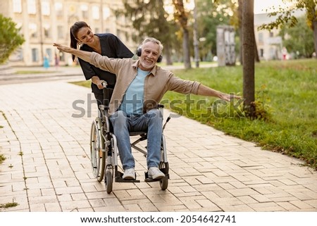 Joyful mature disabled man in wheelchair wearing headphones having fun during a walk in the city assisted by lovely young nurse Royalty-Free Stock Photo #2054642741
