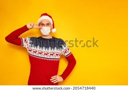 man in New Year's sweater with reindeer, medical mask on his face, Santa Claus hat shows sign of V, peace, victory. isolated yellow background with space for text. concept new year, Christmas, holiday