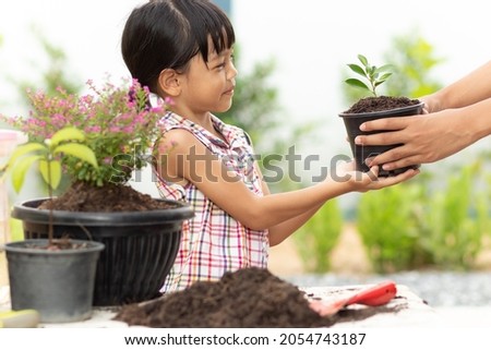 A cute Asian Thai kid girl, aged 4 to 6 years old, is reaching out to pick up a black pot for planting. From the hand of the elder who is extending it to the left of the picture. she so smile happy.