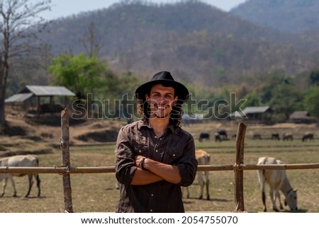 A young hipster is happy and cheerful looks at the camera in front of a cow herd in a cowshed on a dairy farm. Labor, hard work, hope, and an agriculture industry sustainable living of concept ideas.