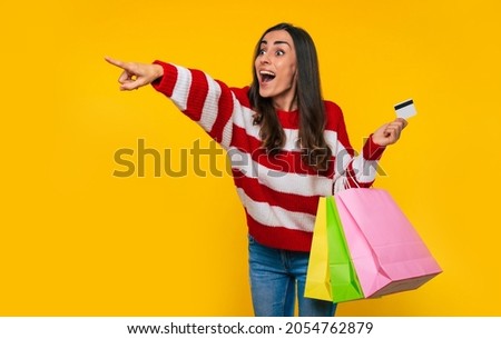 Close up photo of beautiful young happy and excited woman in a striped sweater with many colorful shopping bags and credit card in hands isolated on yellow background