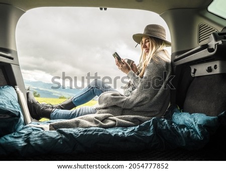 Girl resting in her car with her phone. Woman hiker, hiking backpacker traveler camper in sleeping bag relaxing on top of mountain. Road trip, Health care, authenticity, sense of balance and calmness.