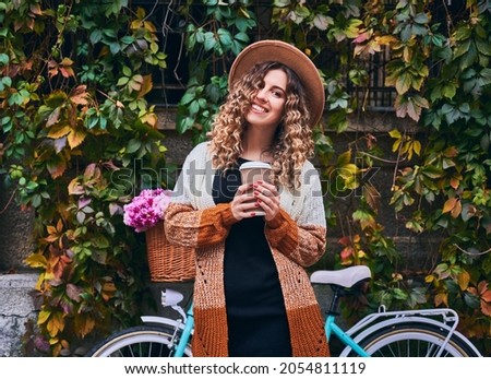 Curly woman posing with vintage bicycle and cup of coffee near green plant wall. Female wearing brown hat and cardigan is travelling by bike with big basket of pink camomiles. Concept of leisure. Royalty-Free Stock Photo #2054811119