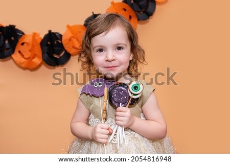 Little girl have fun with Halloween sweets, bat gingerbread cookies, scary eyes, ghosts, lollipops. Making grimaces over isolated background with paper garland of black and orange pumpkins. Copy space