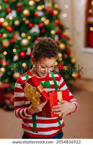 A black boy in a striped red and white jacket holds and open a red box with a Christmas gift, on the Christmas tree background.
