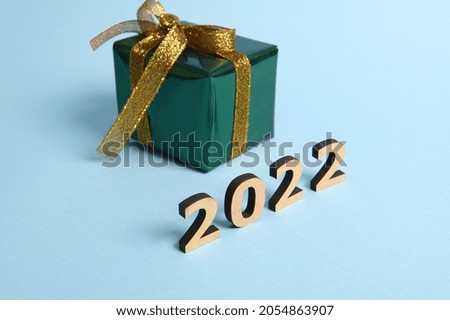 Present box wrapped in shiny glitter green gift wrapping paper and gold ribbon and bow on blue background with wooden numbers year 2022. Christmas, New Year concept. Studio shot for ad with copy space