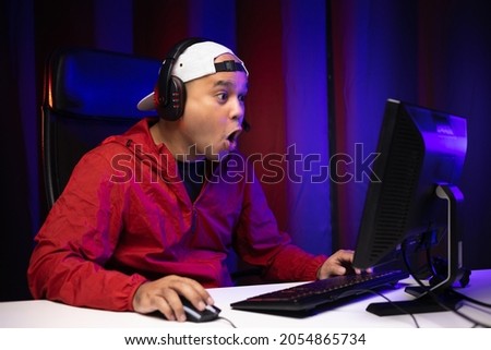 Excited and shocked face of Asian gamer with headphone playing computer pc video game online sitting on chair at living room. Indian professional gamer streaming on social playing game very fun