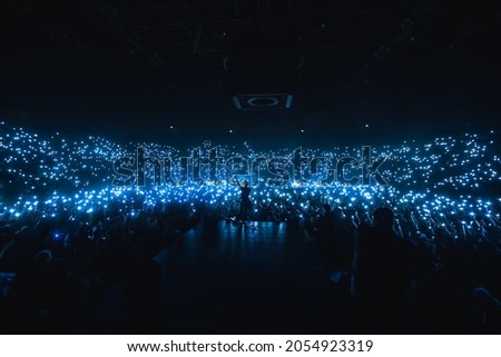 Vocalist in front of crowd on scene in stadium. Bright stage lighting, crowded dance floor. Phone lights at concert. Band blue silhouette crowd. People with cell phone lights. Royalty-Free Stock Photo #2054923319