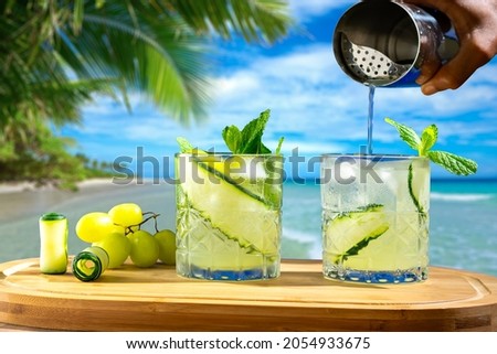two glasses with drinks on a wooden board, in the background a beach, hand holding a cocktail shaker
