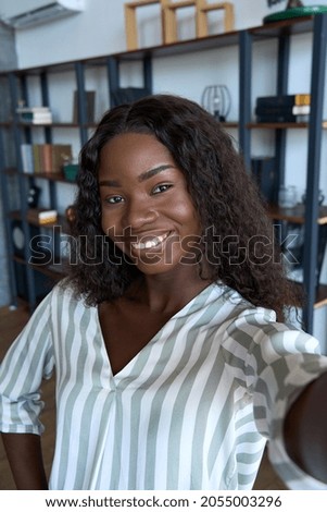 Young happy black African woman holding phone looking at mobile vertical camera streaming vlog, taking selfie portrait, making videocall or recording video on cell at home. Cam view Royalty-Free Stock Photo #2055003296