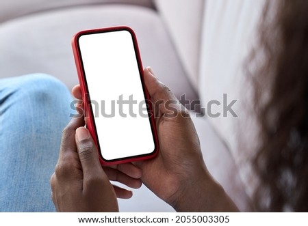 Young black African woman holding smartphone in hand with white black empty mockup screen template using cell phone relaxing on sofa at home. Over shoulder view. Mobile apps advertising concept.