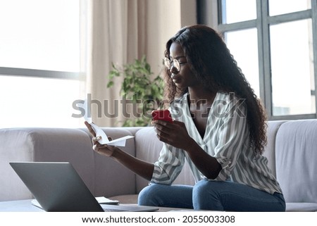 Serious young black African woman holding paper document calculating rent or money savings, paying bills in mobile application on cell phone doing monthly paperwork sitting on couch at home. Royalty-Free Stock Photo #2055003308