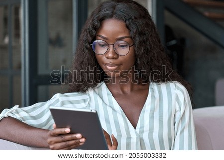 Young black African woman wearing glasses holding digital tablet sitting on couch at home learning online, watching videos, surfing internet, doing e shopping using technology device relaxing on sofa.