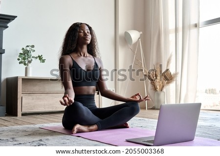 Young calm fit healthy African black woman sitting on floor at home doing yoga breathing exercise, meditating learning online training virtual class on computer. Exercises for mental health concept. Royalty-Free Stock Photo #2055003368