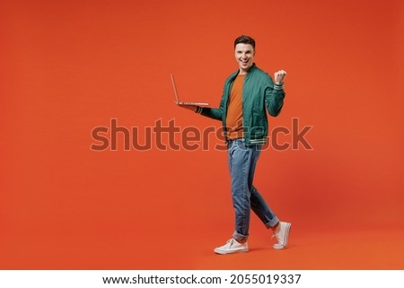 Full size body length vivid happy young brunet man 20s wears red t-shirt green jacket hold use work on laptop pc clenching fists say yes computer isolated on plain orange background studio portrait.