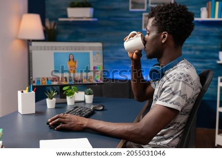 African american man editor drinking coffee while doing photo retouch on computer using creativity post production software. Photographer editing photography sitting at desk in retouching home studio