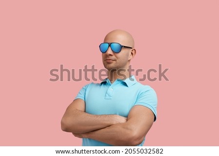 Arrogant bald homosexual man with bristle keeps arms crossed over chest, keeps calm, looks aside, is pleased with choice, gay friendly, wears blue shirt and sunglasses, stands on pink background