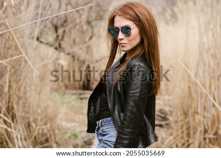 Portrait of a girl with long hair in a leather jacket and sunglasses, in the autumn forest. Young hipster or rocker woman posing on the street. 
