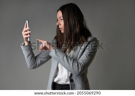 businesswoman dressed in a jacket and trousers in a casual style uses smartphone for work and contact with partners, studio portrait on gray background, Use touch screen, take photo or use video call