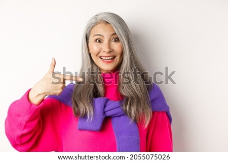 Close up of beautiful and stylish asian senior woman smiling, pointing at herself, showing logo, standing over white background
