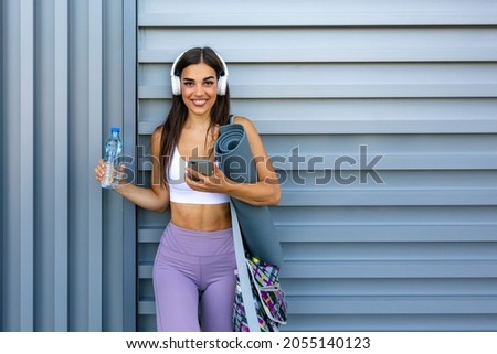 Sportswoman in headphones holding a plastic bottle. Shot of beautiful female runner standing outdoors holding water bottle. Fitness woman taking a break after running workout.