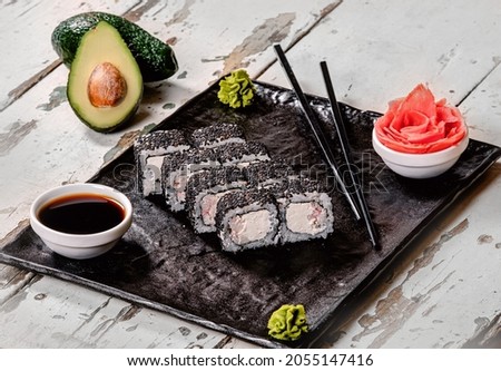 Roll sushi on a white vintage table, in a black plate (Sashimi bun), sliced avocado in the background, a white bowl (seuzarus) with soy sauce on the side, sticks (hasi and saibashi), wasabi and ginger