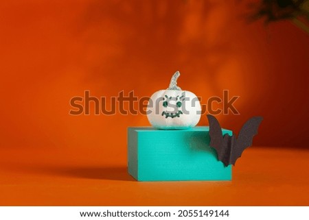 White pumpkin with face on podium over orange background with shadows and copy space. Holiday Halloween party concept.
