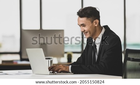 Positive Asian male entrepreneur sitting at table and browsing netbook while working on project in workplace and looking laptop computer screen.